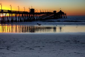 View of the pier from the beach as the sun sets behind and to the left of the pier and seagulls dot the shoreline.