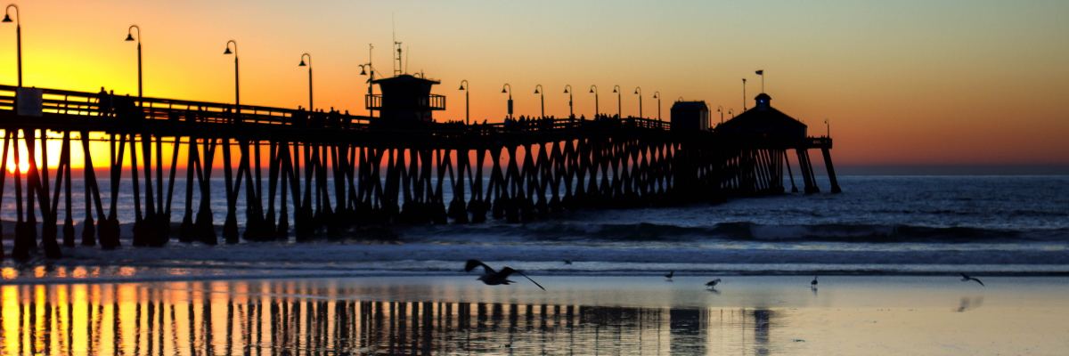 View of the pier from the beach as the sun sets behind and to the left of the pier and seagulls dot the shoreline.