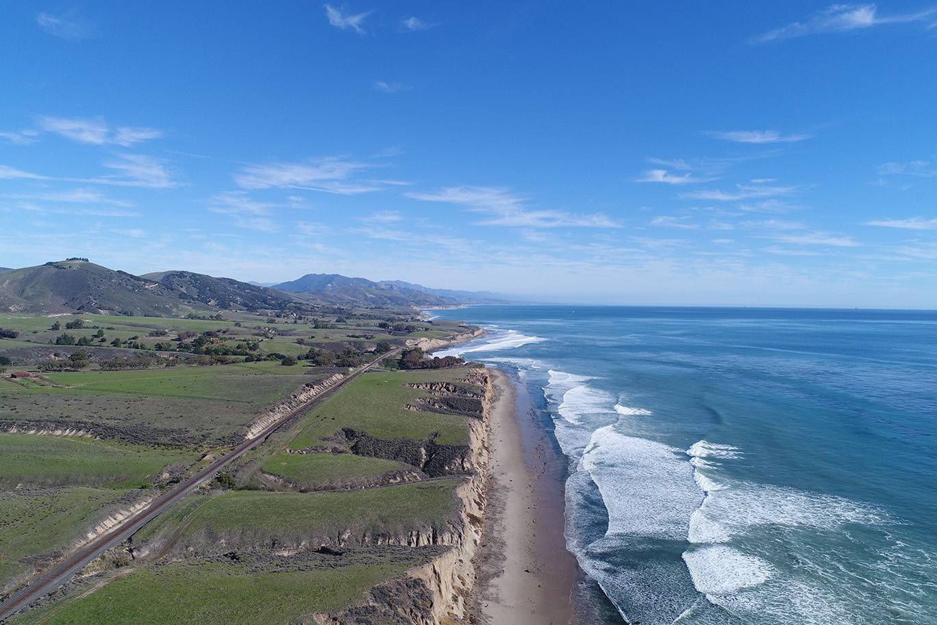 drone image of Hollister Ranch with the empty beaches and green hills to the left and breaking waves and the open Pacific ocean to the right.