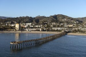 looking onto the pier from the water with the beach, city and hills in the background and a clear sky overhead.